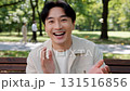 Cheerful Asian man laughing and waving hello while sitting on park bench in summer day outdoors 131516856