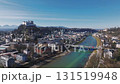 Smooth ascending drone view of Salzburg, Austria, featuring Hohensalzburg Fortress, the Salzach River, tree lined pathways, and urban greenery. 131519948