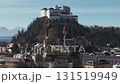 Aerial view of Hohensalzburg Fortress in Salzburg, Austria, with rolling hills, scattered buildings, and snow capped Alps in the background. 131519949