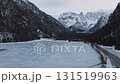 Aerial view of a frozen lake in Dolomites, Italy, with snow capped peaks, dense evergreen forests, and a narrow pathway along the lake's edge. 131519963