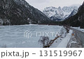 Sweeping aerial view of a frozen lake in Dolomites, Italy, surrounded by snow covered peaks, sparse trees, and a winding road along the icy surface. 131519967