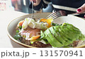 Woman's hands using a knife and fork to cut a poached egg on a healthy toast with avocado, smoked salmon, and lettuce. The bright yellow yolk beautifully spills over the dish 131570941