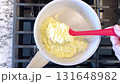 Hand holds a red spatula, stirring bright scrambled eggs in a non-stick frying pan, the blue flame of a gas stove underneath ensuring a warm, hearty meal. 131648982