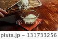Close-up of a chef's hands transferring chopped fresh white onions, coarsely sliced into rings, from a wooden cutting board over a wooden table into a special bowl. 131733256