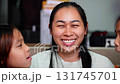 A joyful Asian mother smiling while her two daughters kiss her on both cheeks at home. The image represents family love, affection, motherhood, happiness, and bonding between parent and children.  131745701