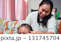 A warm and candid moment of a mother braiding her daughter's hair while the little girl smiles brightly at home. 131745704