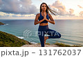 Black woman with braids practicing yoga tree pose on cliff overlooking ocean during beautiful sunset 131762037