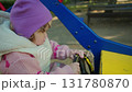 Child plays on a red playground toy in a sunny park surrounded by trees and benches 131780870