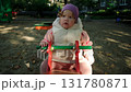 Child plays on a red playground toy in a sunny park surrounded by trees and benches 131780871