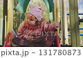 A happy toddler in a pink sweater slides down a playground slide, laughing with joy. The setting features green trees and colorful play equipment, showing a typical day at the park. 131780873