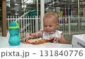 A girl with short hair bites into a pizza, tearing off a large piece with both hands. She eats with concentration, chews, and looks at the water in a sippy cup on the table with interest. 131840776