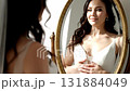 A stunning bride-to-be, with dark, wavy hair and a white slip dress, looks at her glowing reflection in an ornate golden mirror 131884049