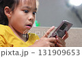 Close-up of an Asian child playing a mobile game on smartphone at home, showing modern lifestyle, technology use, and digital entertainment among kids today. 131909653