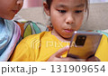 Two Asian girls sitting on sofa watching smartphone screen together at home, representing childhood, learning, technology, and digital lifestyle in modern family life. 131909654