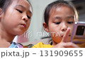 Two Asian girls sitting on sofa watching smartphone screen together at home, representing childhood, learning, technology, and digital lifestyle in modern family life. 131909655