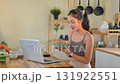 Morning wellness routine showing woman in sportswear eating yogurt bowl beside a laptop and fresh ingredients 131922551