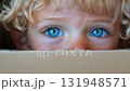 A cute child with beautiful blue eyes peeking out from a cardboard box, displaying curiosity and innocence. 131948571