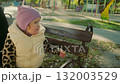 A little child sits on a park bench, wearing a warm vest and a hat. She curiously inspects flower petals while enjoying the sunny autumn day in a vibrant playground. 132003529