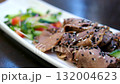 Serving of sliced boiled beef tongue with pickled cucumbers and bell peppers drizzled with olive oil soy sauce and crushed garlic topped with black and white sesame and microgreens on white plate. 132004623