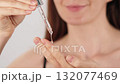 Close-up of an unrecognizable woman holding a pipette with facial serum. A drop of transparent oil or gel drips from the glass dropper. Skincare, cosmetic product, liquid collagen, beauty routine. 132077469