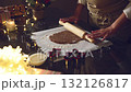 Close-up of hands rolling out gingerbread cookie dough with a rolling pin, surrounded by twinkling Christmas lights and decorations. Perfect for holiday baking and festive moments. 132126817