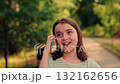 smiling girl talking on smartphone outdoors in sunny green park., happy child making phone call while walking along tree-lined path., cheerful tween chatting on mobile phone during warm summer 132162656