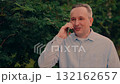 enjoying conversation amidst verdant tree scenery., holding smartphone close, man enjoying conversation, green trees creating soothing backdrop., strolling through leafy garden., content man speaking 132162657