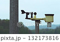 Small weather station with anemometer and rain gauge is rotating in the wind, mounted on a metal pole in front of a blurred background of green trees and hills 132173816