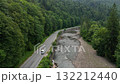 Aerial view of a white car driving on an asphalt road winding along a shallow mountain river through dense forest 132212440