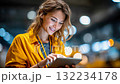 Smiling woman in a yellow jacket using a tablet in an industrial setting, representing technology, logistics, and workplace efficiency 132234178