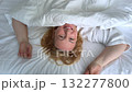 Happy mature woman lying on bed under blanket, smiling upside down at camera and making heart shape with hands 132277800