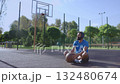 Positive athletic bearded African American male basketball player sitting on outdoor court, wearing wireless headphones and enjoying music while relaxing after streetball training. Positive athletic bearded African American male basketball player sitting on outdoor court, wearing wireless headphones and enjoying music while relaxing after streetball training. 132480674