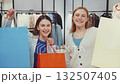 Two smiling women holding colorful shopping bags after successful shopping trip, enjoying retail therapy in modern clothing store 132507405