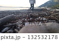 A woman approaches a large log on the beach and sits down. In the spring, a girl in a sports jacket and baseball cap has come to the beach to sit and think 132532178