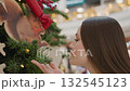 Close-up of young lady smiling as she smells decorative Christmas tree with a sign of satisfaction, background features festive decorative lights creating a joyful, holiday shopping atmosphere 132545123