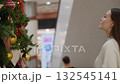 Young woman standing in front of Christmas tree, looking excited and amazed with bokeh light effect in background, shoppers walking by and building inscription visible in vibrant mall 132545141