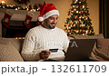 Man in festive sweater and Santa hat smiles while shopping online with credit card, cozy holiday atmosphere, camera zooms in on joyful expression 132611709