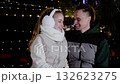 Festive winter evening showcasing young couple laughing, illuminated by soft holiday lights, sharing intimate moment outdoors 132623275