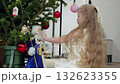 Young blond girl decorating christmas tree with glass ornaments, positioning santa claus figurine among sparkling branches, generating warm holiday ambiance 132623355