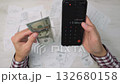 Hands holding cash and a smartphone with a calculator app, with numerous receipts scattered on a desk. This image represents personal finance management and digital money tracking 132680158