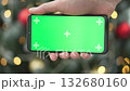 A hand holds a modern smartphone with a vibrant green screen against a blurred backdrop of twinkling Christmas lights and colorful ornaments, capturing the joyful holiday atmosphere 132680160