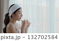 Beautiful young asian woman with headband enjoying tea standing near window with satisfied. 132702584