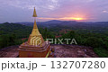 A stunning aerial view of a golden pagoda nestled amidst lush green hills during a vibrant sunset, with dramatic clouds and soft sunlight casting a serene glow over the landscape. 132707280