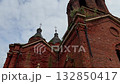 Low angle shot features historic red brick church with ornate towers and weathered crosses silhouetted against cloudy sky, intricate facades and wild grass on ledges evoke sense of faded grandeur and 132850417