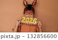 A shy child dressed up as a horse with a painted face and ears on his head hides behind paper numbers 2026. 132856600