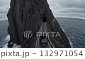 Car traveling on a narrow road carved into a steep cliffside, approaching a tunnel entrance with the vast, moody ocean stretching out under a dramatic sky 132971054