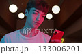 Young Trendy Guy in White T-Shirt Using his Modern Smartphone while Standing at Futuristic Neon Club Lights Background. Communication and Technology Concept. Close Up 133006201