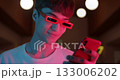 Young Trendy Guy in Stylish Modern Party Glasses Stand Using his Smartphone, Looking to Screen and Typing at Futuristic Neon Club Lights Close Up shot 133006202