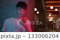 Young Stylish Millenial Boy in White T-Shirt Using his Smartphone for Chatting, Typing Messages while Standing at Futuristic Neon Club Lights Background Close Up Shot 133006204