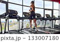 Sofia, Bulgaria - 11.10.2025. A young woman in sportswear on a treadmill adjusts the settings and starts running in a modern gym with large panoramic windows and a city view. Real time video. 133007180